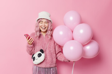 Happy Asian girl laughs joyfully expresses positive emotions holds smartphone and inflated balloons prepares for celebration of special occasion dressed in winter outerwear poses against pink wall