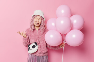 Upset dejected woman has bad mood celebrates birthday alone holds bunch of inflated balloons dressed in outerwear expresses negative emotions isolated over pink background. Spoiled holiday concept