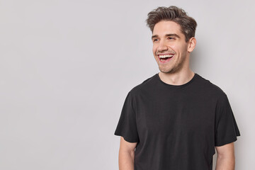 Horizontal shot of handsome cheerful man looks happily away smiles broadly wears casual black t shirt isolated over white background with blank copy space being in good mood laughs at something