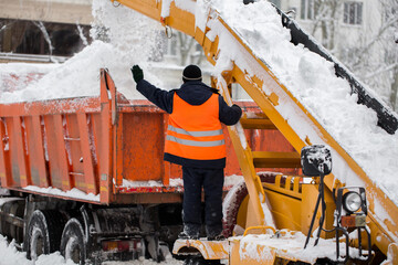 Claw loader vehicle removes snow from the road.  Uniformed community service worker helps load snow into a truck.