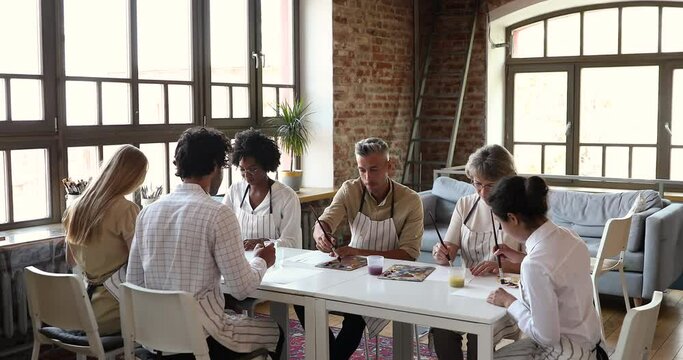 Smiling mature female painter psychologist hold art therapy lesson for young people of some ethnic groups. Happy diverse fine art school students sit at desk learn to draw lead by aged lady professor