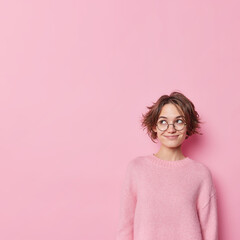 Indoor shot of beautiful dreamy woman with short dark hair smiles gently and looks away wears round...