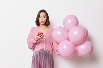 Obraz na płótnie Canvas Shocked young Asian woman holds mobile phone gets message holds bunch of inflated balloons celebrates anniversary dressed in festive clothes isolated over white background. Celebration concept
