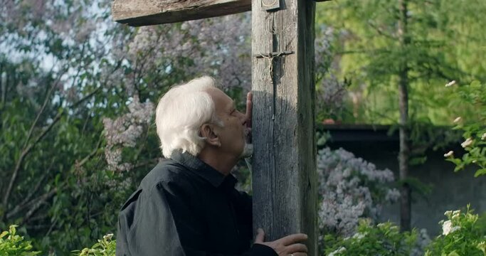 Sad old gray haired man stands near big old wooden cross at the cemetery. Sorrow and sadness about lost of close people. Religious man, 4k 60p Prores