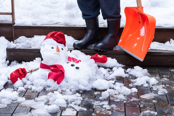 Unhappy snowman in mittens, red scarf and cap is melting  outdoors on wet pavement and man’s boots and shovel near it. Approaching spring, warm winter, climate change concept  