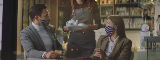Female waiter in cafe bringing ordered beverages of table. Man and woman talking together and enjoying evening. Wearing protection face masks. - Powered by Adobe