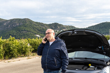 Caucasian young adult calling the roadside assistance service on the phone, with a broken down car behind.