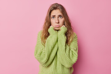 Sad young European woman with long hair has innocent pleading expression keeps hands under chin purses lips wears knitted green sweater isolated over pink background complains about something