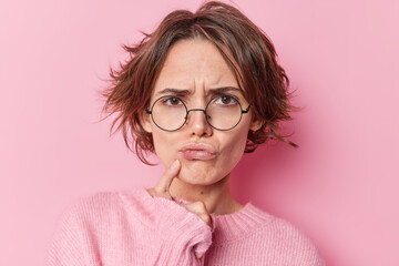 Serious European woman tries to find decision thinks hard keeps finger near lips feels displeased has gloomy expression wears round spectacles long sleeved jumper isolated over pink background - obrazy, fototapety, plakaty