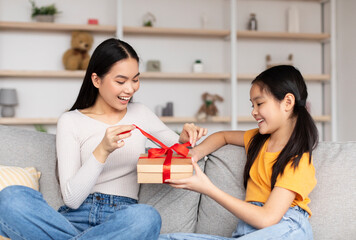 Happy teenage girl congratulates young asian lady and gives box with present, sit on sofa