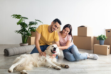 Positive young diverse couple stroking their dog in new home on moving day, full length. Home...