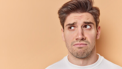 Obraz premium Studio shot of unhappy brunet young man has sulking expressionn looks sadly away feels disappointed has some problems isolated over brown background with blank space for promotional content.