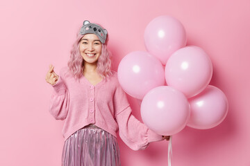 Obraz na płótnie Canvas Happy positive Asian woman makes korean like sign shapes mini heart expresses love holds bunch of balloons wears sleepmask jumper and skirt celebratess birthday isolated over pink background