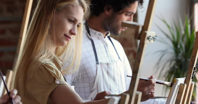 Young persons of diverse gender race smiling European woman focused Hispanic man take exam at private drawing school painting in oils on canvases. Multiethnic students learning academic course of arts