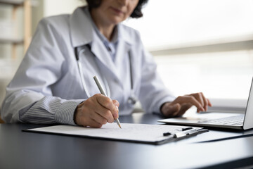 Close up older female general practitioner doctor handwriting notes on clipboard, web surfing information online on computer, managing patient's appointment or writing healthcare prescriptions.