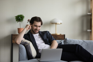 Happy relaxed millennial 30s man looking at computer screen, enjoying using software applications, communicating distantly by video call, spending time shopping online, lying on comfortable sofa.