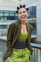 Vertical shot of happy fashionable woman dressed in stylish clothes keeps hands in pockets laughs...