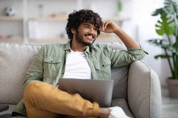 Happy indian guy in casual sitting on couch with laptop
