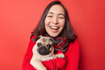 Joyful beautiful woman has eastern appearance poses with pug dog enjoys spending free time with favorite pet expresses positive emotions stands indoor against vivid red background. Animals concept