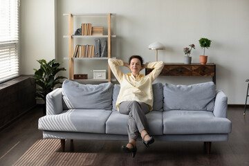 Obraz premium Happy peaceful middle aged senior retired woman relaxing on cozy sofa with closed eyes, breathing fresh air, enjoying carefree leisure weekend time in modern living room, sleeping alone at home.