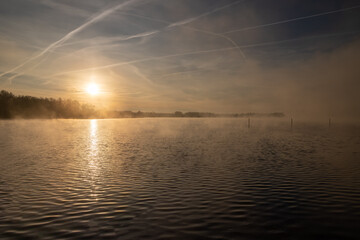 Early winter morning at a lake during a sunrise and the sunlight reflects over the water of the lake and creates a fairy tale landscape together with the frozen ground and grass and a veil of mist