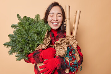 Positive Asian woman holds bouquet of spruce branches paper rollers and retro garland for...