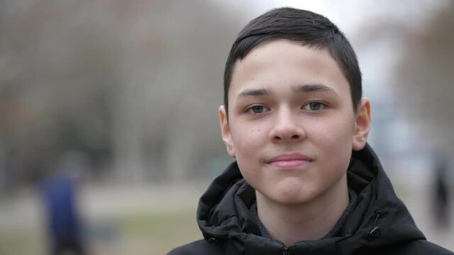 Portrait of a 13-year-old European boy dressed in a black jacket. He stands on the street in autumn and looks at the camera against a blurred background. Slow motion, close-up, shallow depth of field