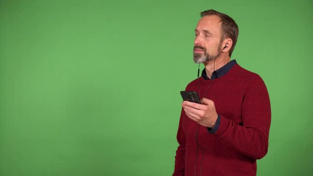 A middle-aged handsome Caucasian man listens to music with earphones on and a smartphone with a smile - green screen background