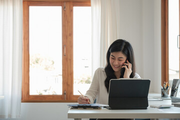 Happy business asian manageress working at her desk in the office taking a call on her mobile phone while writing notes on a notepad