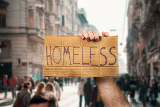 A man hold a cardboard sign with the inscription HOMELESS. Defocused background with street and people. Hands close-up. The concept of helping vagabonds