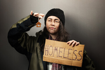 A beggar smiling woman holds a cardboard sign with the inscription homeless and looks at a keychain...