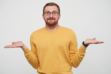 Studio portrait of young bearded male student wears yellow sweater holding copy space on both palms, looking into camera with confused facial expression, isolated over white background