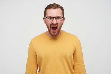 Studio portrait of young bearded male student wears yellow sweater screaming into camera with angry facial expression, isolated over white background