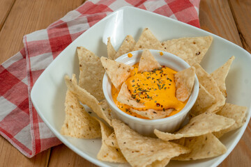 Nachos dipped in cheddar on a bowl on a wooden table. Under the dish there is a red and white kitchen cloth and the background is grey. Vertical image