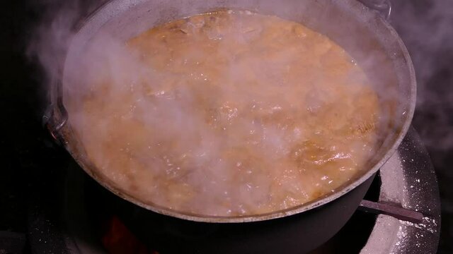 Goulash is prepared with seasoning and meat. Steam from a hot dish. Kitchen in a cauldron. Stir with a kitchen spatula. Campfire food.