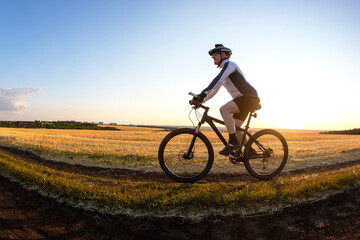 Fototapeta na wymiar The cyclist rides a bike on the road near the field against the backdrop of the setting sun. Outdoor sports. Healthy lifestyle.