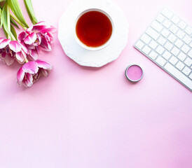 Fototapeta na wymiar Cup of black tea with bouquet pink peony tulips and white wireless keyboard, lips balm on pink background