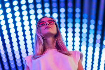 Teen hipster girl in stylish glasses standing on blue tunnel with neon light wall background,...