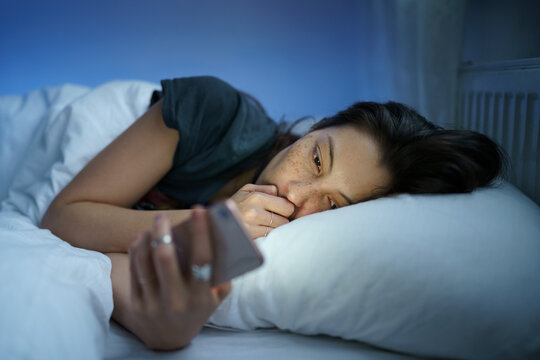 Depressed sleepless young woman follow cheating ex-boyfriend in social media lying tired in bed and suffering from insomnia. Anxious female with sleep disorder look at smartphone screen in dark room