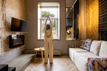 Relaxed woman meditating at modern apartment, looking at window. Concept of mental health and home comfort. Caucasian woman wearing beige sleepwear. Wide interior view