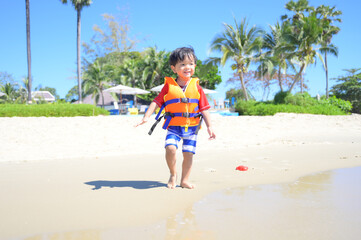 Asian baby boy with swimwear and life vest walking on the tropical beach .