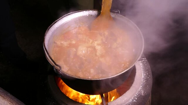 Goulash is prepared with seasoning and meat. Steam from a hot dish. Kitchen in a cauldron. Stir with a kitchen spatula. Campfire food.