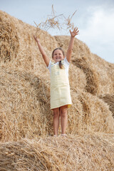 Naklejka na ściany i meble Portrait of blissful small girl smiling with teeth standing on high haystack throwing up hay and looking at it wearing sundress. Having fun away from city on field full of golden hay.