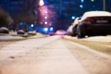 Winter in the night city. Sidewalk in the yard. Residential area. Snowy road. Parked cars. Working lights. Juicy colors. Focus on snow. Close up view from road level.