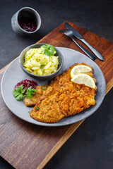 Traditional deep-fried schnitzel with potato salad, cranberries and lemon slices served as close-up on a modern design plate on a wooden board
