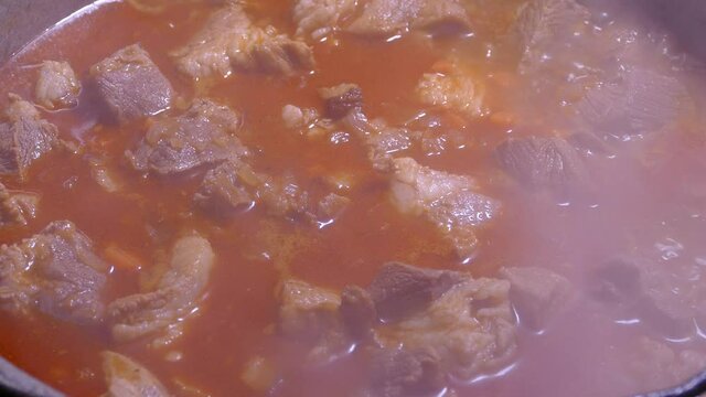 Goulash is prepared with seasoning and meat. Steam from a hot dish. Kitchen in a cauldron. Stir with a kitchen spatula. Campfire food.