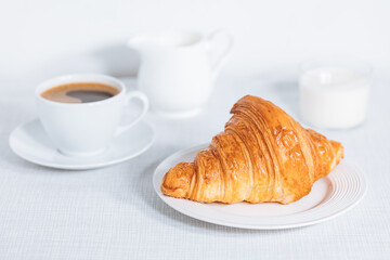 Croissant in bright white environment, with cup of coffee and milk and other croissant in blurry background. Simple and elegant breakfast setting and scene.