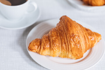 Croissant in bright white environment, with cup of coffee and milk and other croissant in blurry background. Simple and elegant breakfast setting and scene.