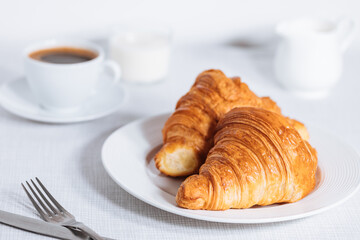 Croissant in bright white environment, with cup of coffee and milk and other croissant in blurry background. Simple and elegant breakfast setting and scene.
