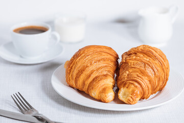 Croissant in bright white environment, with cup of coffee and milk and other croissant in blurry background. Simple and elegant breakfast setting and scene.